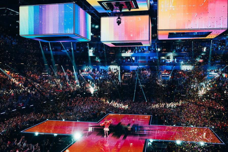Künstler steht auf einer großen ESC-Bühne in Kreuzform, umgeben von Konfetti, LED-Würfeln über der Bühne und einem voll besetzten Publikum während einer Live-Show.©Sarah Louise Bennett_EBU_38609 by SRGSSR